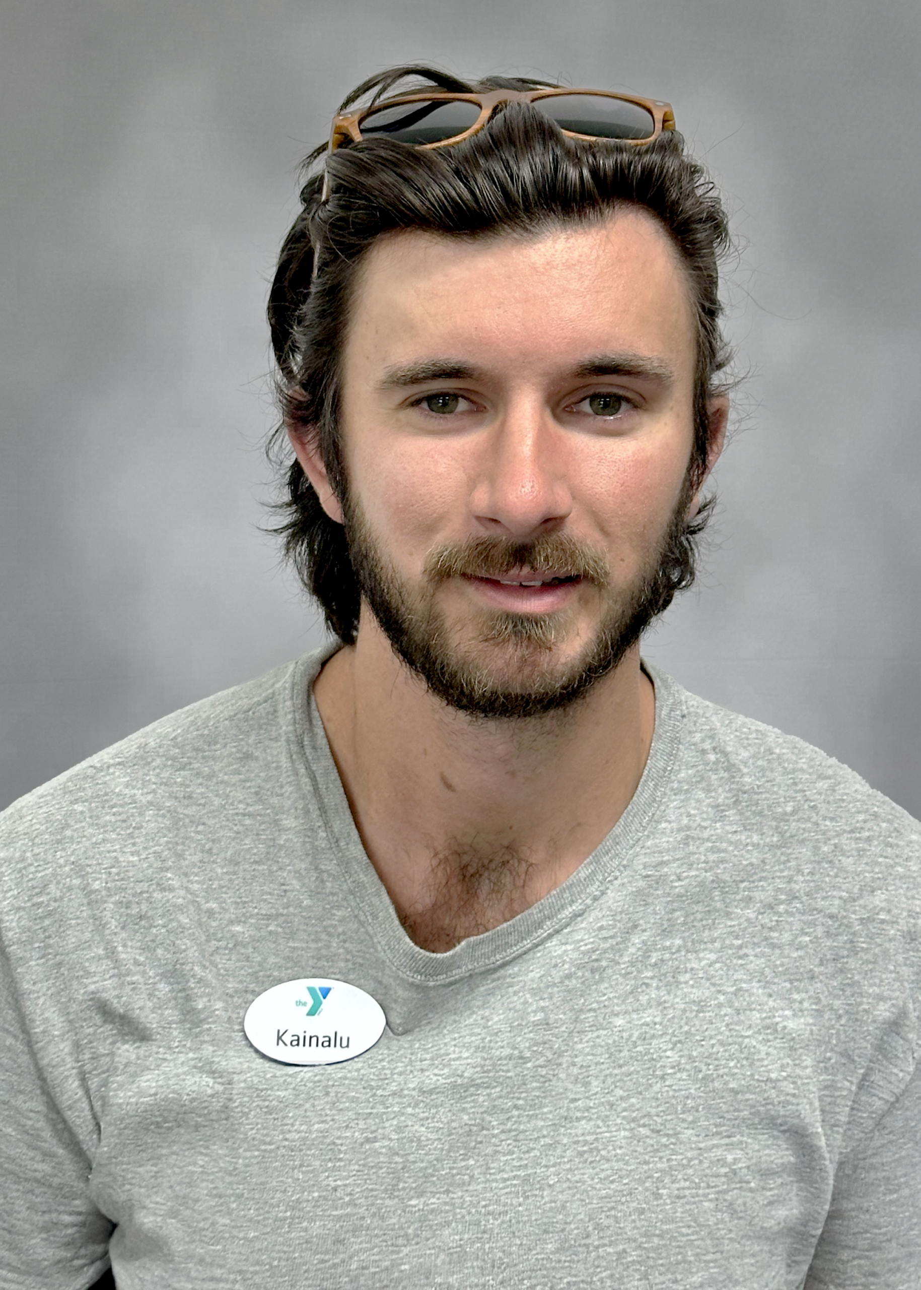 Portrait of a man with dark hair and a beard, wearing a gray shirt and sunglasses on his head; badge reads 'Kainalu'.